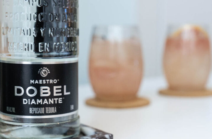 Close-up of a "Maestro Dobel" liquor bottle with two pink cocktail drinks out of focus in the background.