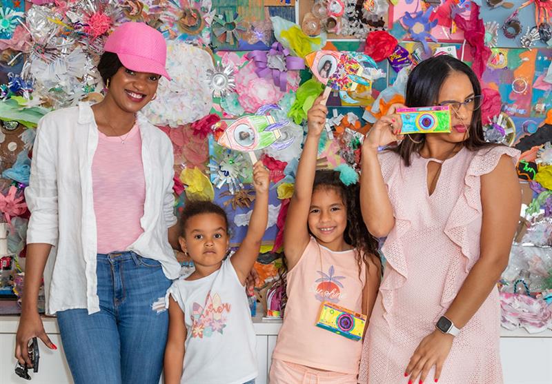 Mothers and daughters pose holding disposable cameras in front of recycled artwork at The Bass Creativity Center Family Day.